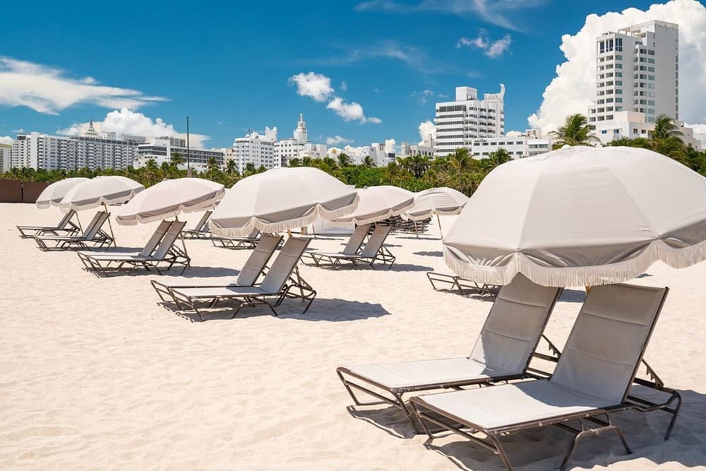 White loungers and umbrellas on the sand at the private beach club, with the Miami Beach skyline behind